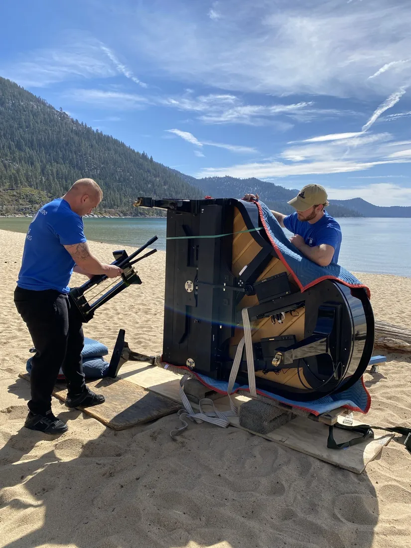 Crew setting up a grand piano on a lakeside beach.
