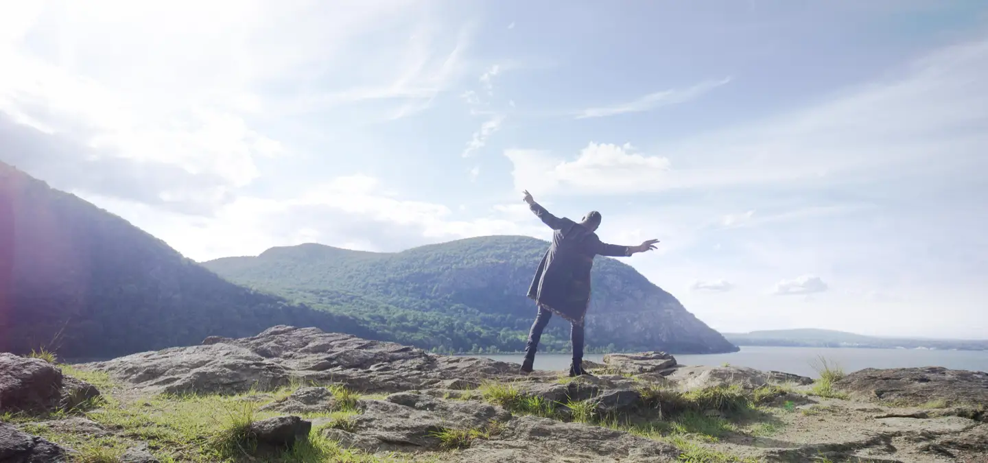 Music video still: a performer with arms spread wide on a mountaintop above a river valley
