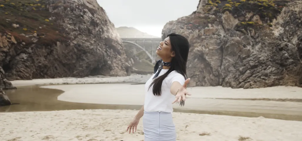 Music video still: the singer with arms open on the beach below Bixby Creek Bridge in Big Sur, in Ana Beatriz’s “Ver Você”