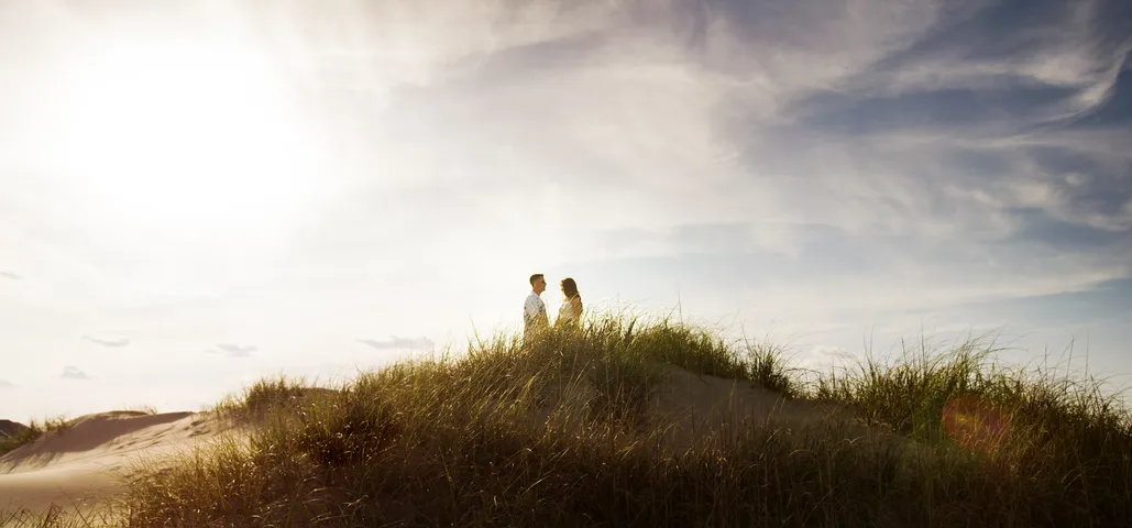 Music video still: a couple standing on top of a grassy dune under a big sky in Celeste Kellogg’s “There’s a Beach Somewhere”