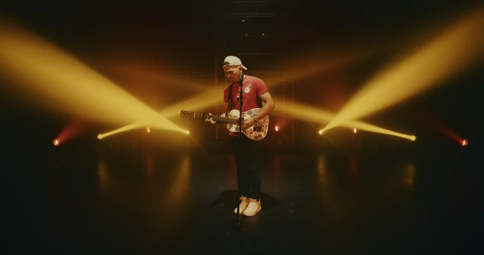 Music video still filmed at WRECK’D Productions in Tinton Falls, NJ: a singer in a red shirt and white cap playing acoustic guitar under amber beams