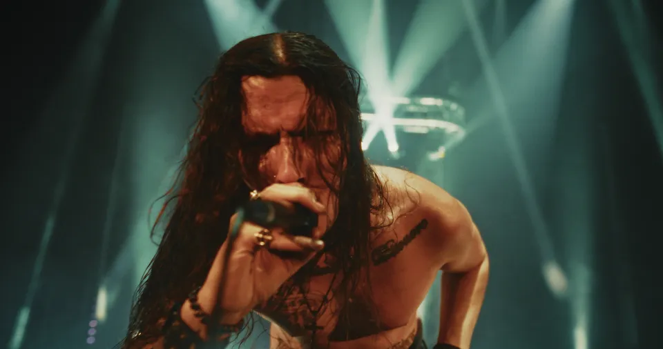 Music video still: a long-haired singer screaming into the mic under green and white beams