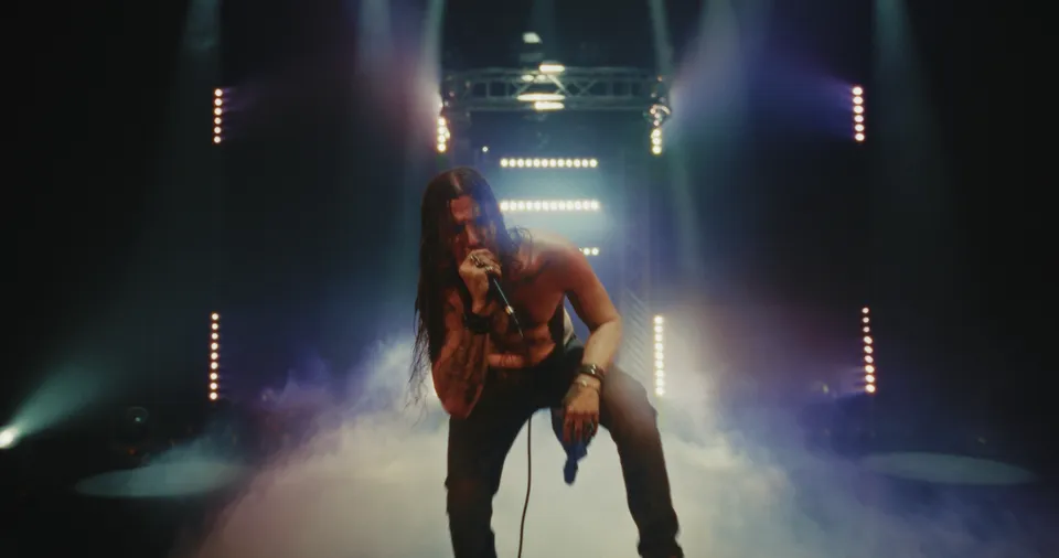 Music video still filmed at WRECK’D Productions in Tinton Falls, NJ: a long-haired singer crouched at the mic in stage haze under white light bars