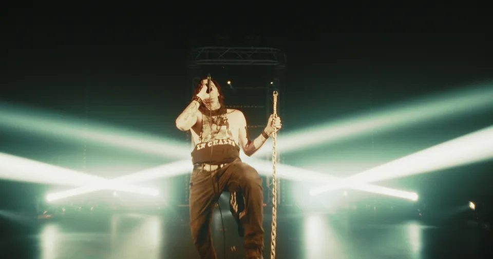 Music video still: a shirtless singer sitting with a chain under crossing light beams
